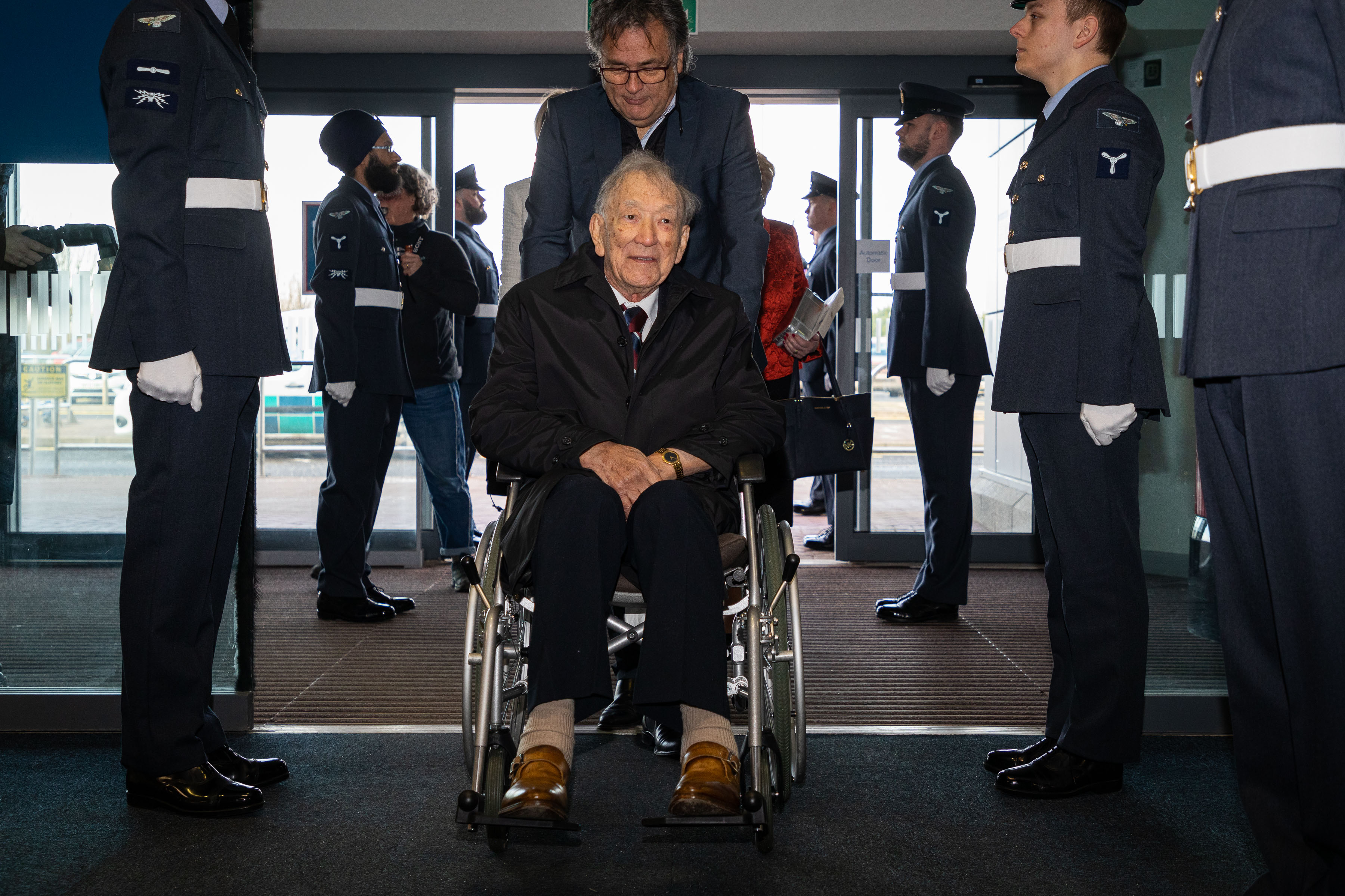 Image shows RAF aviators to attention as veteran is wheeled between them.